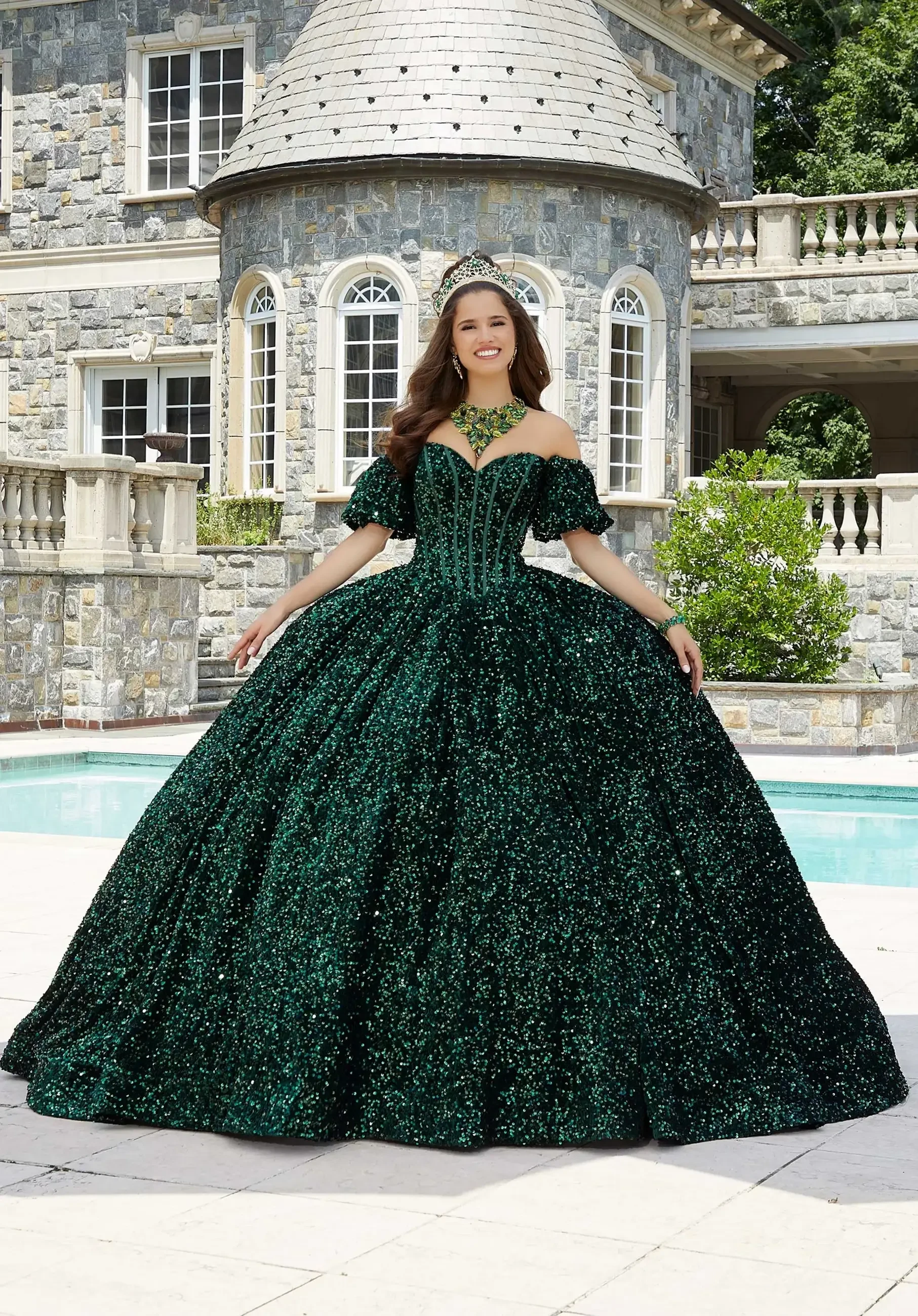 A woman wearing a large, elegant green ball gown stands in front of a stone mansion with an outdoor pool.