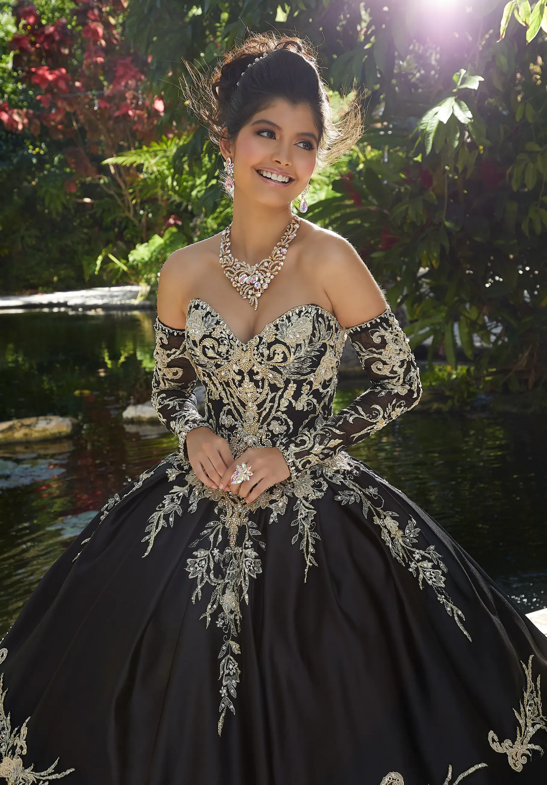 A smiling woman in an intricately designed black ball gown with gold embroidery and off-shoulder sleeves, wearing a statement necklace and rings, posed elegantly outdoors near water surrounded by greenery.
