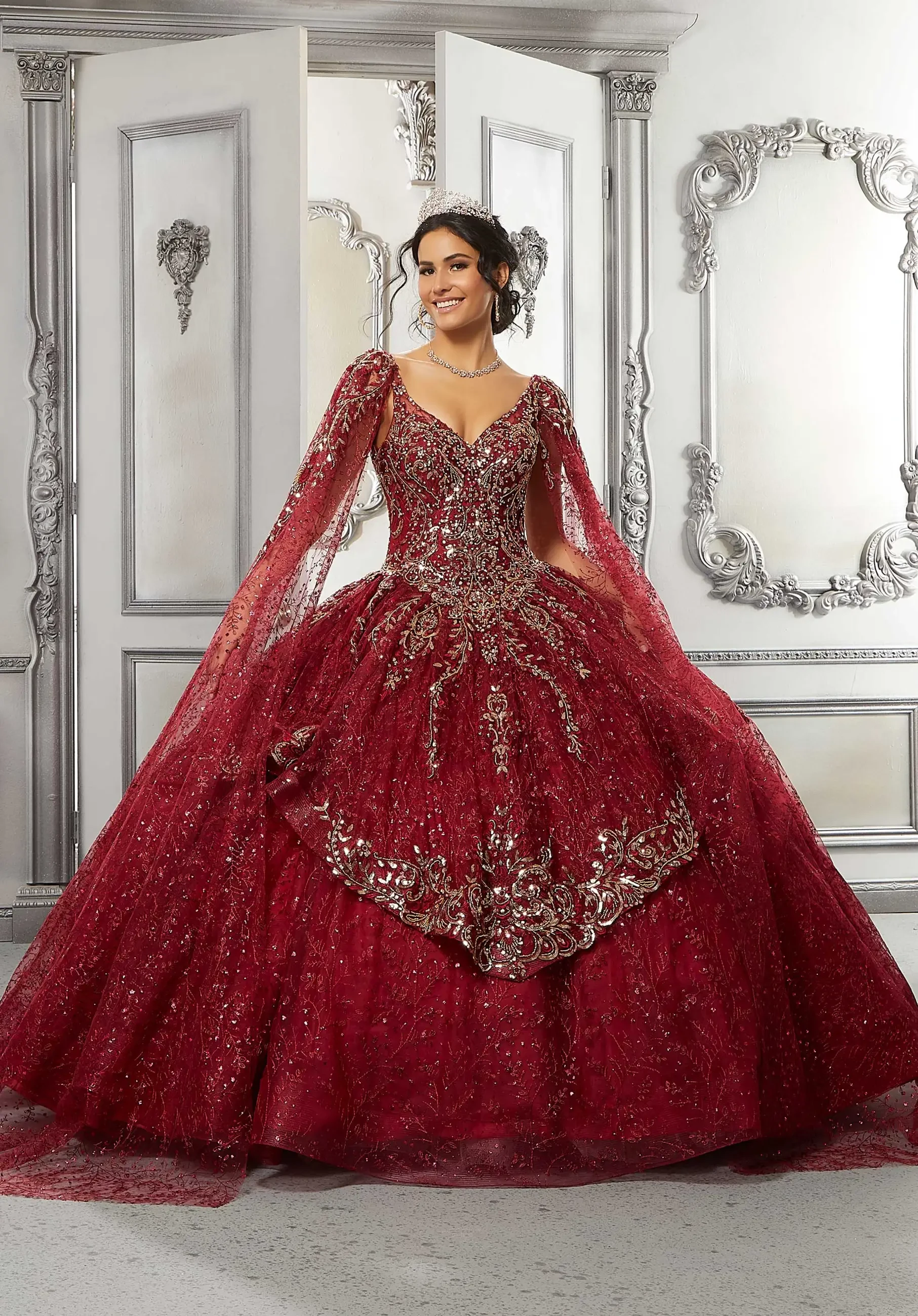 A woman wearing an elegant, deep red gown with intricate beadwork and a flowing train, standing in a lavishly decorated room with ornate mirrors and doorways.