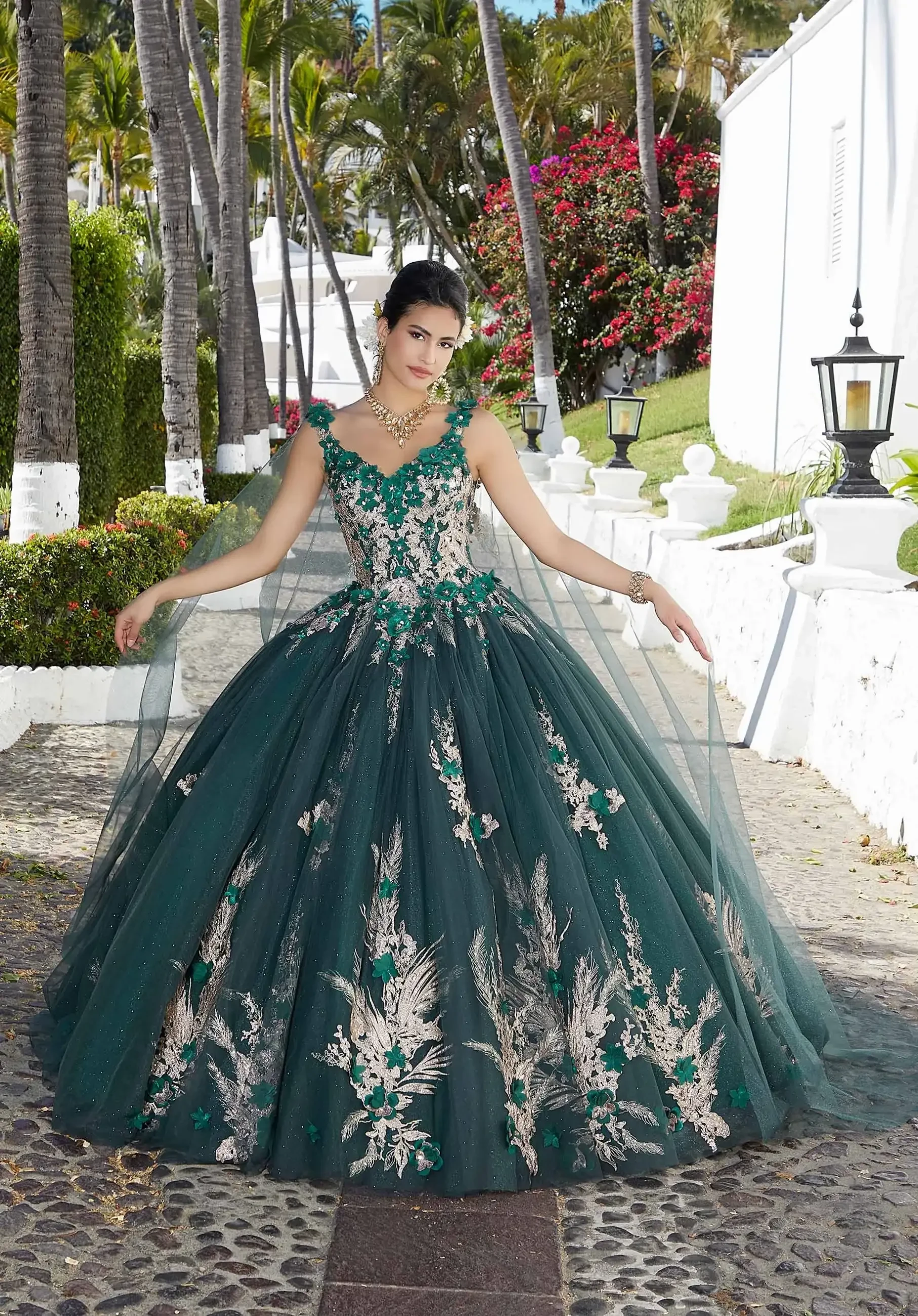 A young woman in a stunning green ball gown with intricate floral embroidery, standing outdoors surrounded by palm trees and white buildings.