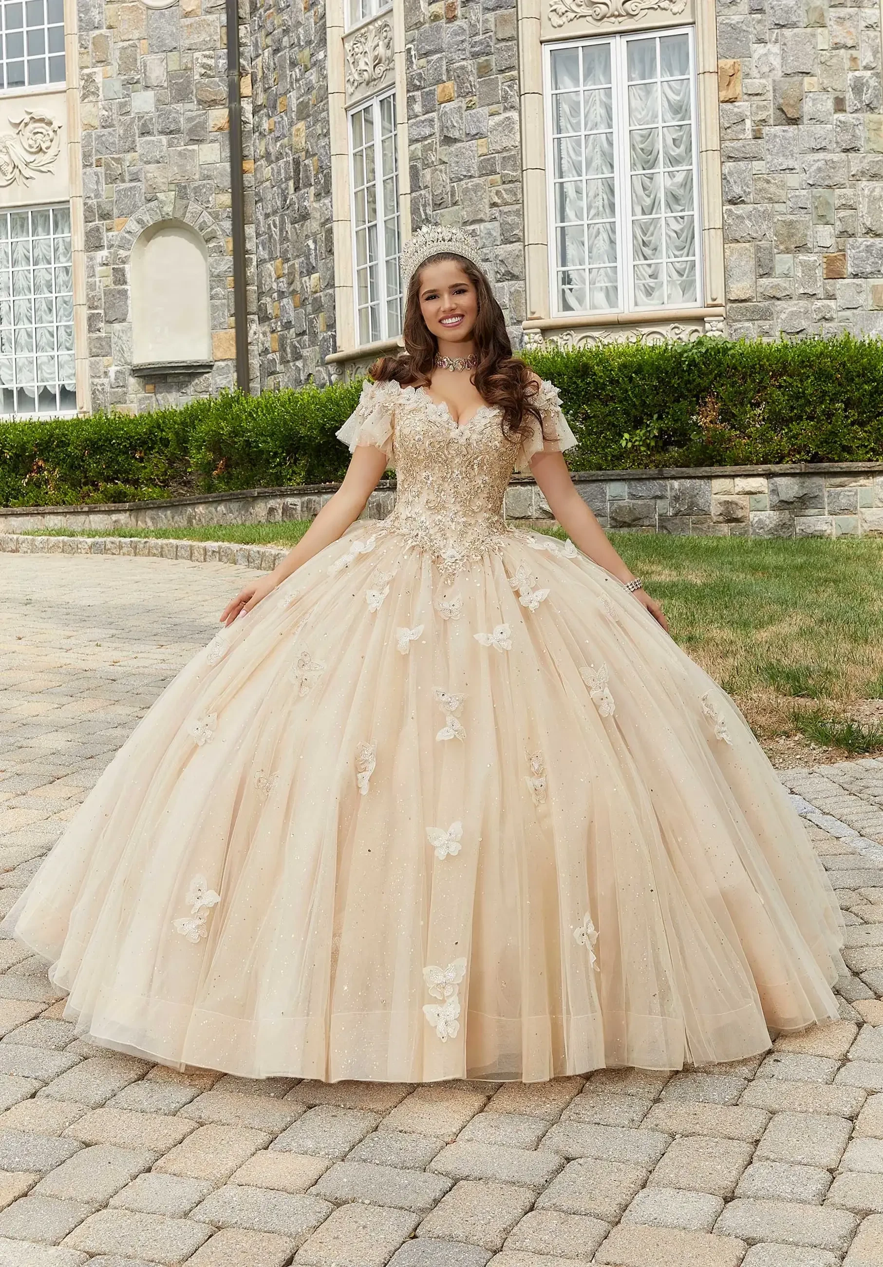 A woman wearing a beautifully detailed, ball gown style wedding dress with floral embellishments, standing gracefully on a stone pathway in front of an elegant building.