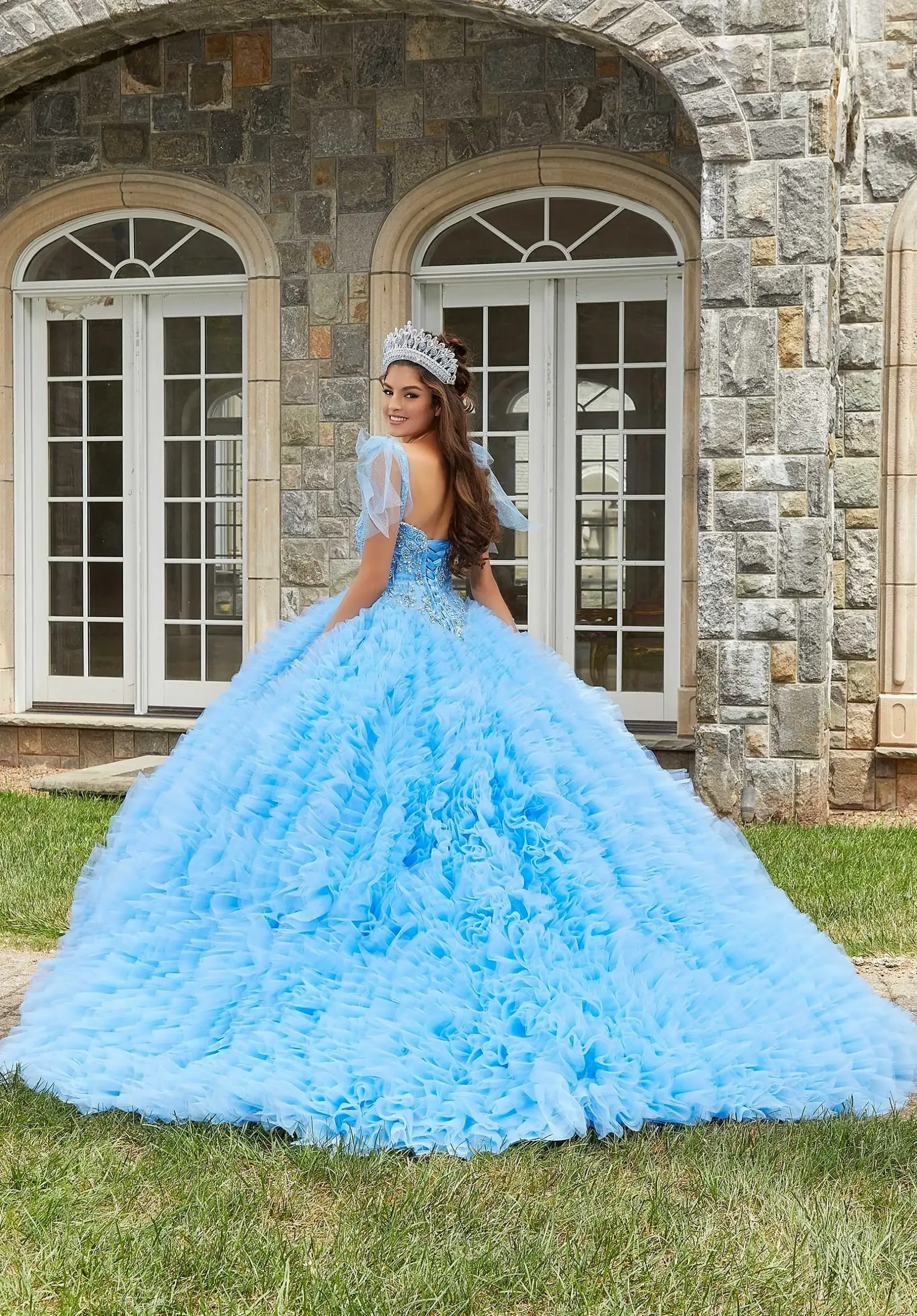 A person wearing a large, fluffy blue gown with a tiara, standing with their back to the camera in front of a stone building.