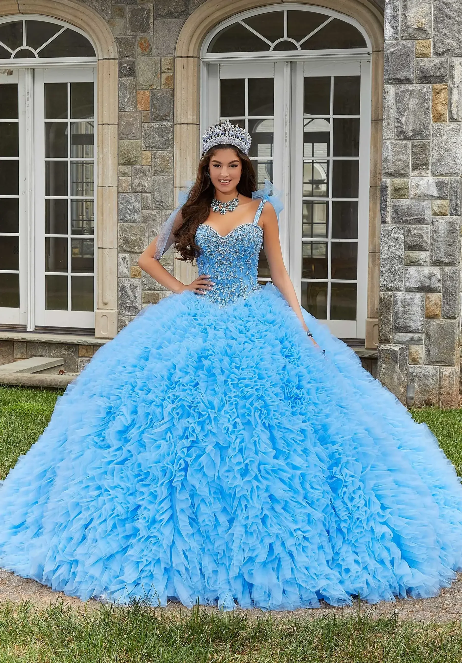 A young woman in a large, blue, ruffled ball gown stands outside a stone building, wearing a tiara and jewelry.