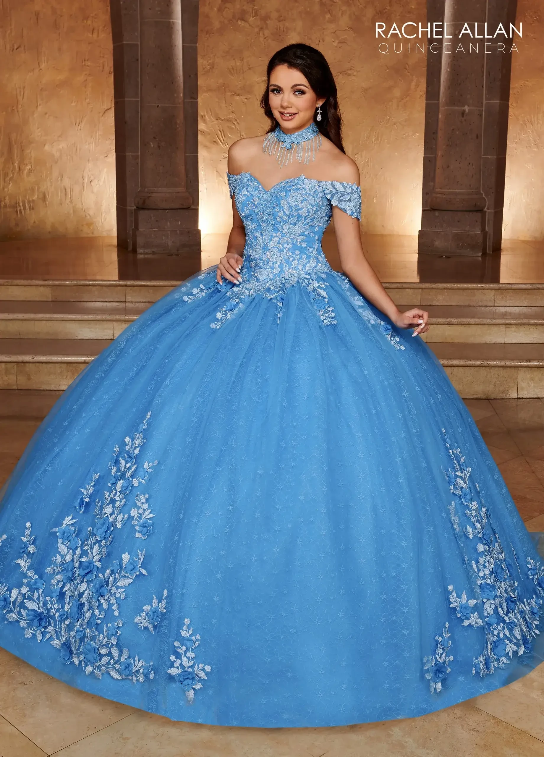 A young woman wearing a large, light blue ball gown adorned with floral embellishments, standing in an elegant indoor setting. The gown features off-the-shoulder sleeves and a fitted bodice.