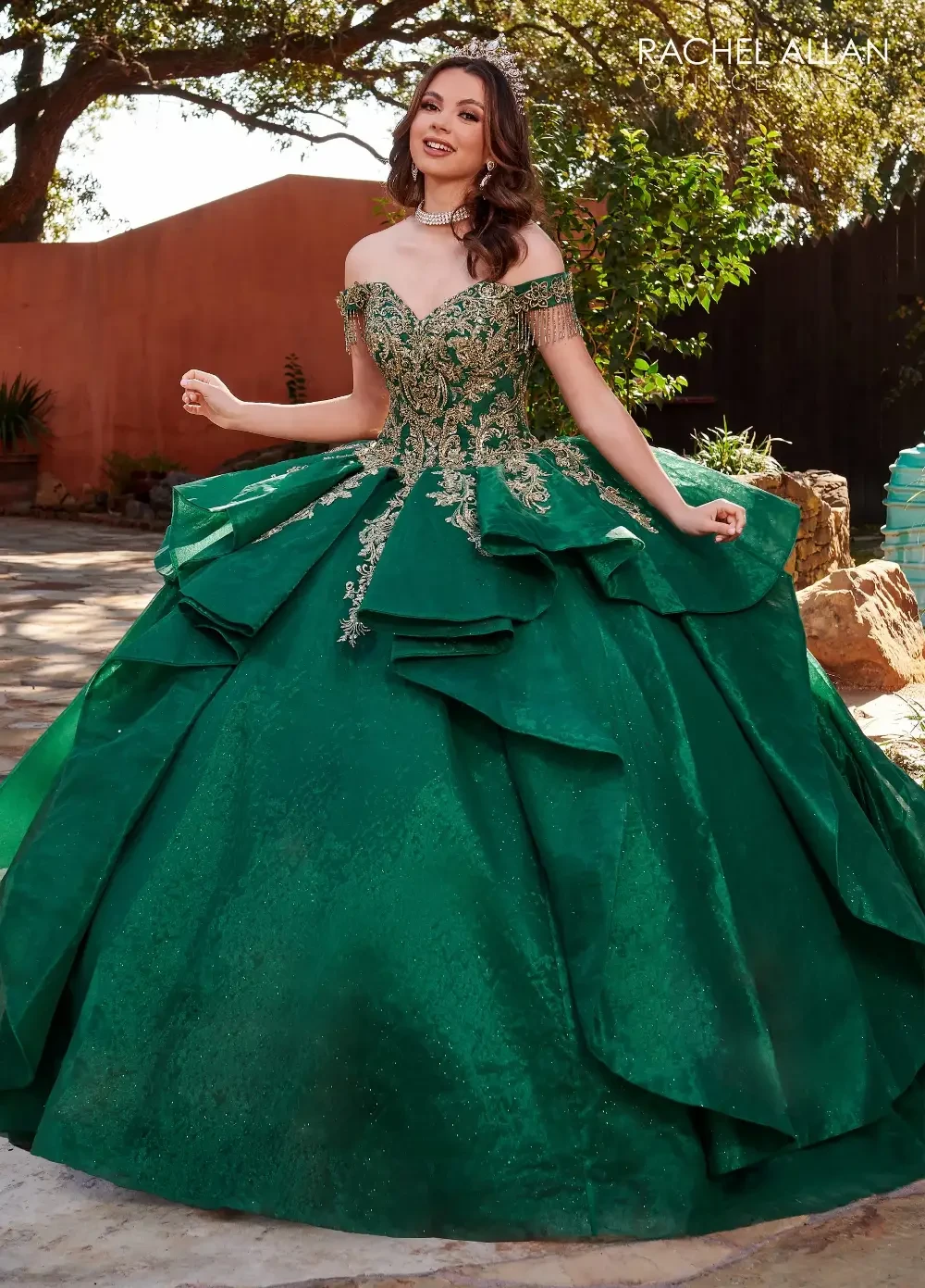 A young woman wearing a detailed green ball gown with off-the-shoulder sleeves and layered ruffles, standing in an outdoor setting.