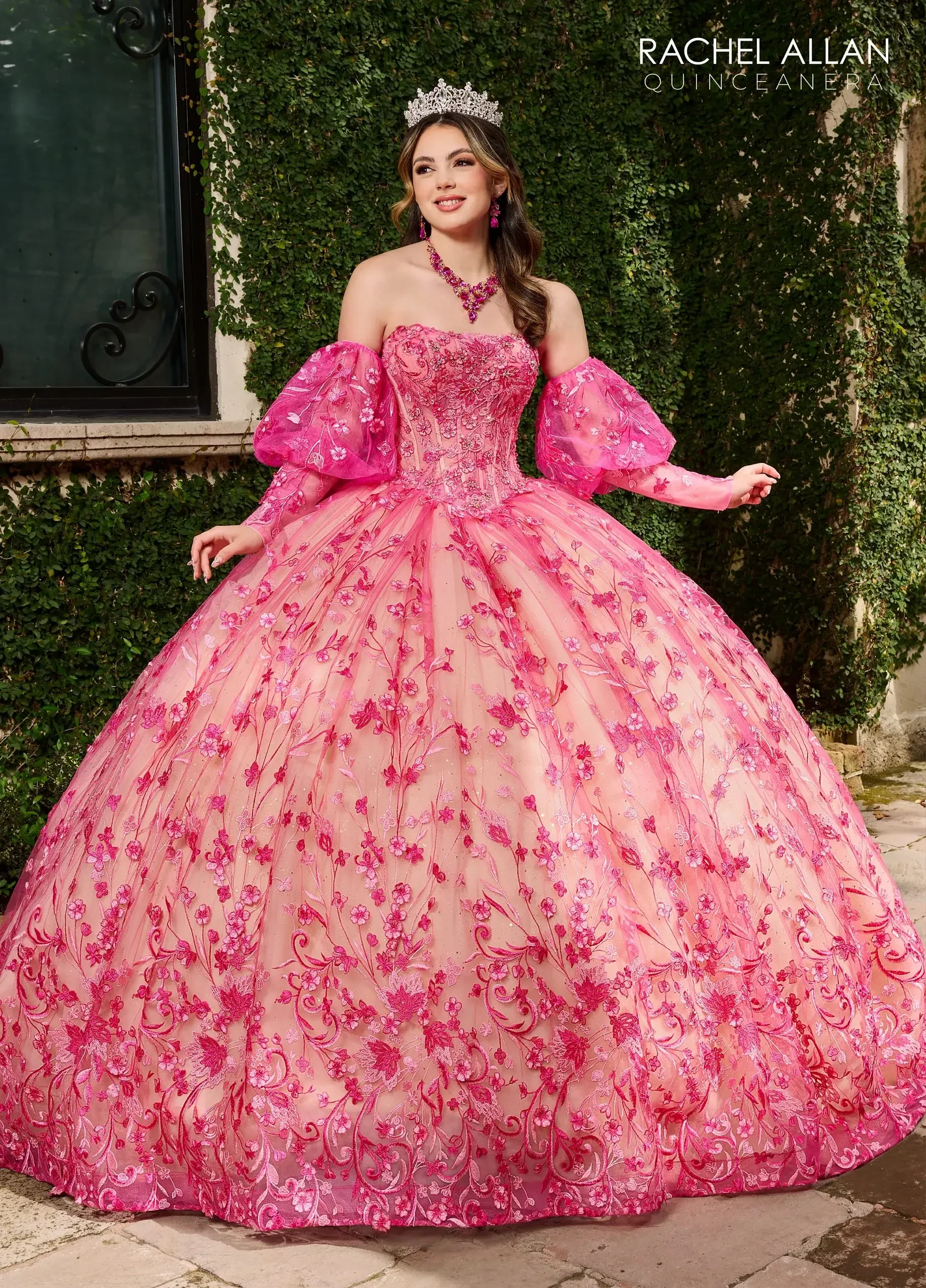 A young woman in a pink ball gown with floral decorations and puffed sleeves, wearing a tiara and jewelry, standing outdoors near greenery.