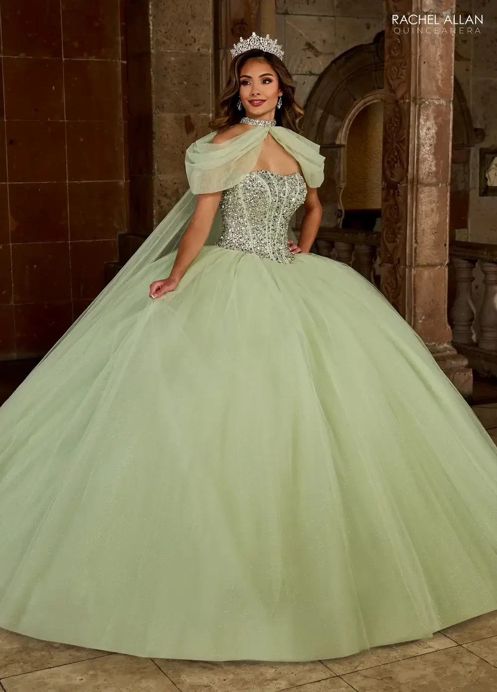 A woman in a light green ball gown with a fitted bodice and a full skirt, wearing a tiara and standing in front of a decorative stone background.