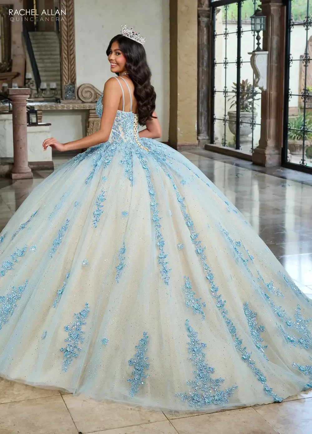A woman in a luxurious, light blue ball gown with intricate embellishments, standing in an elegant indoor setting.