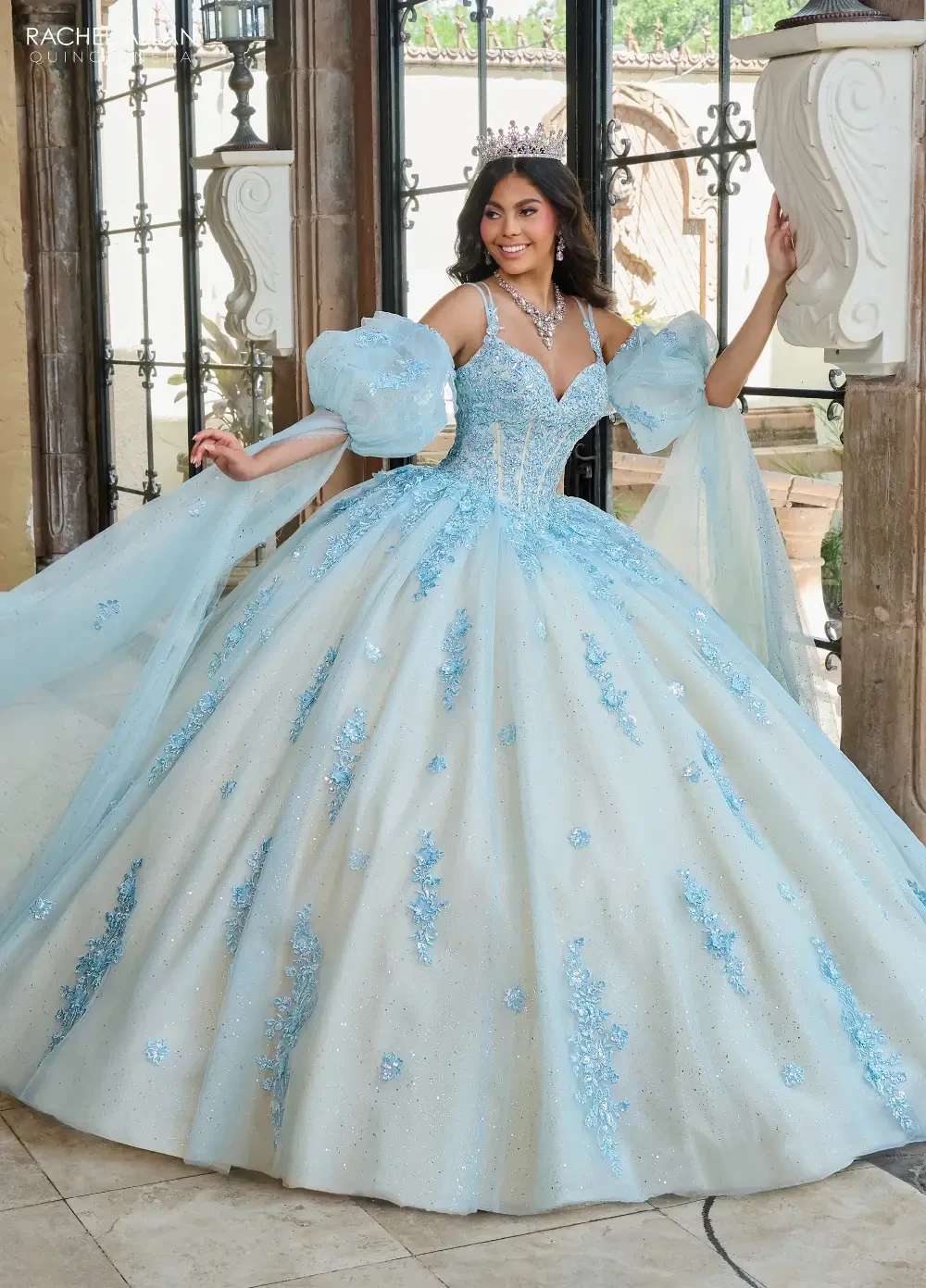 A young woman in a stunning light blue ball gown with intricate beadwork and puffed sleeves, standing gracefully in an elegant indoor setting.