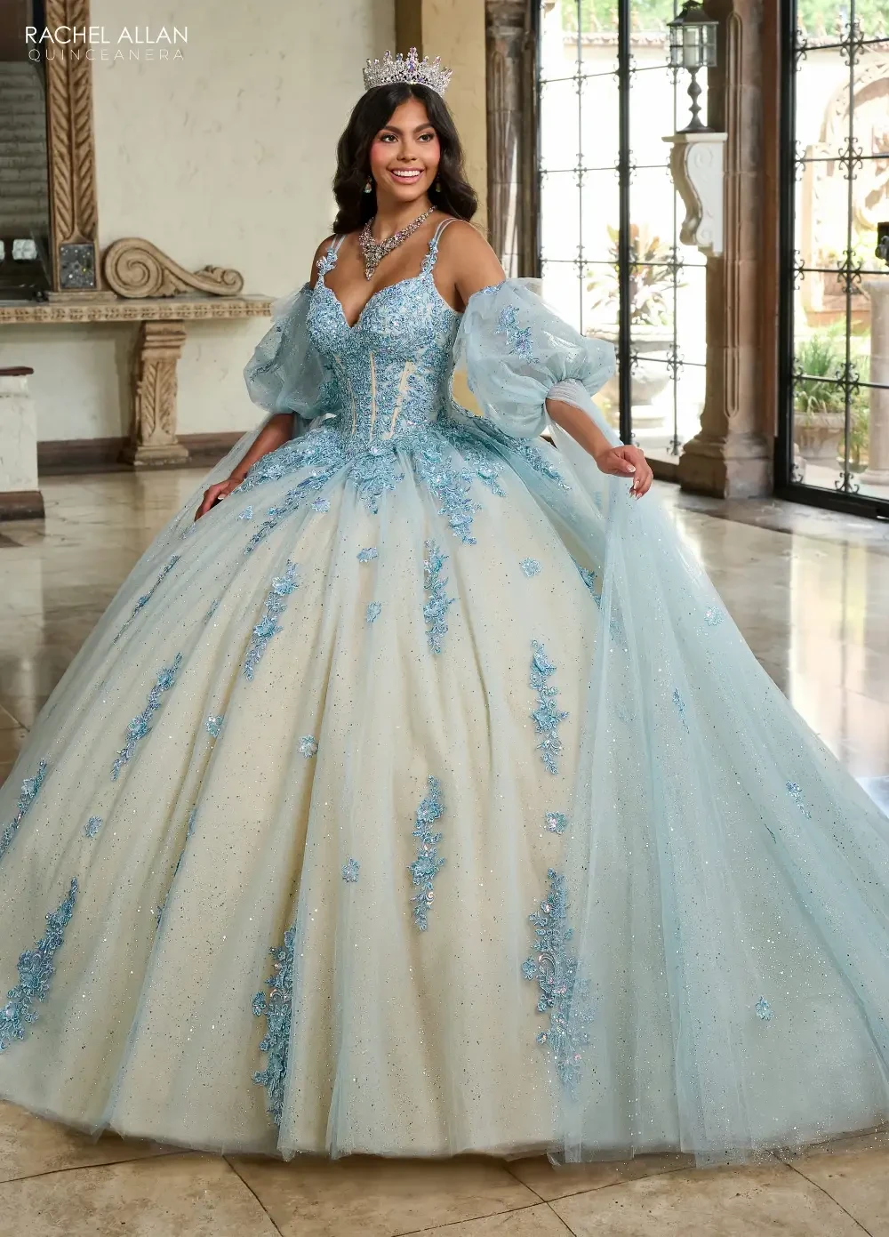 A woman in a luxurious, light blue ball gown with intricate embellishments, standing in an elegant indoor setting.