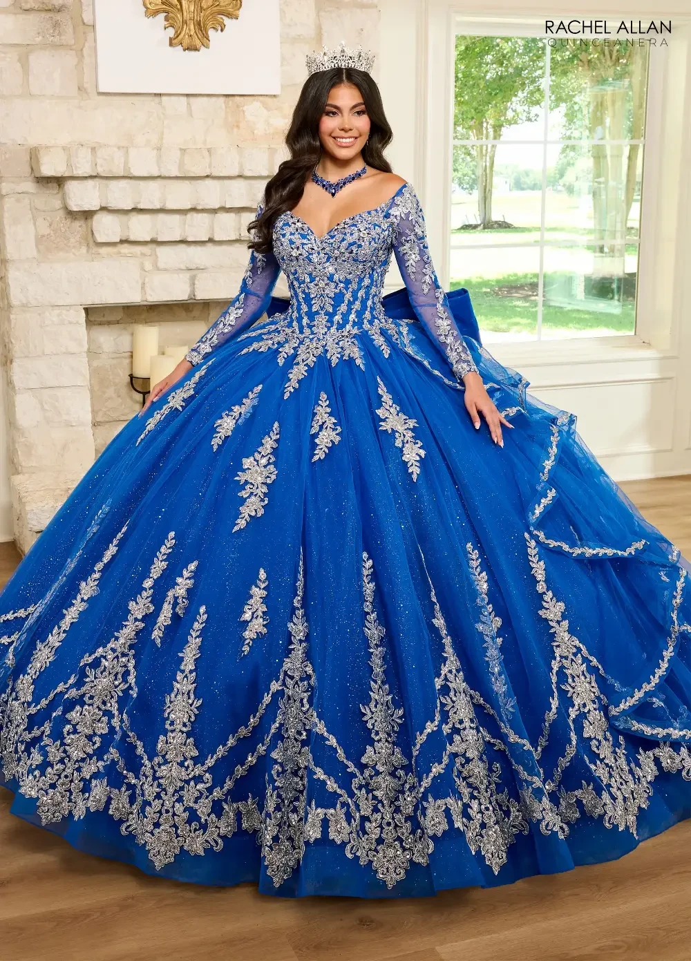 A young woman wearing a large, beautiful blue ball gown adorned with intricate silver designs, standing in a brightly lit room with a stone fireplace. She has long dark hair and is wearing a tiara and matching jewelry.